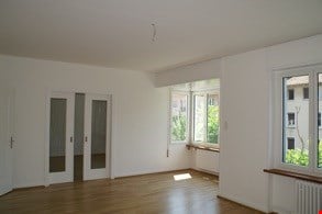 Empty room with wooden flooring, white walls, large windows, and a sliding glass door.