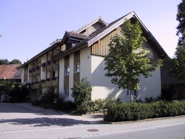 Two-story residential building with a pitched roof, beige facade, and wooden paneling on the upper level. Balconies with flower boxes are visible on the left side.