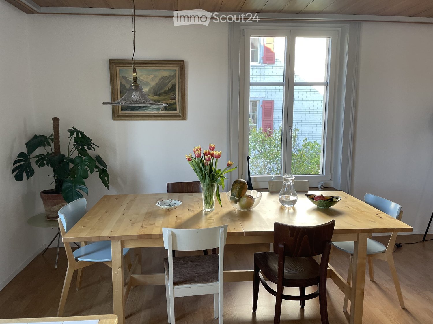 Bright dining area with a wooden table, multiple chairs, a vase with tulips, and decorative bowls. Large windows provide natural light, and a potted plant adds greenery.