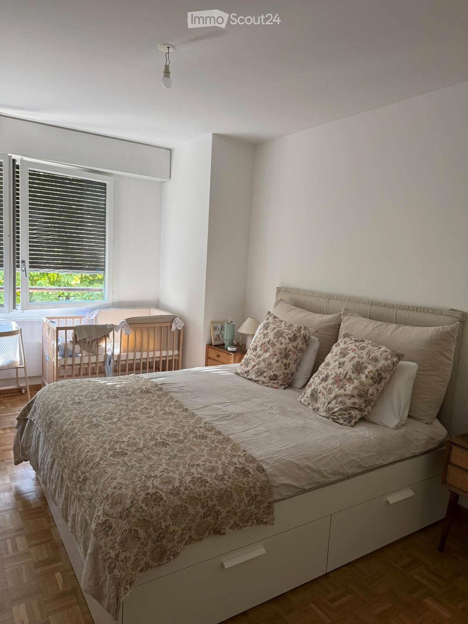 A well-lit bedroom with a large bed, nightstands, a cradle, and a chair near the window.