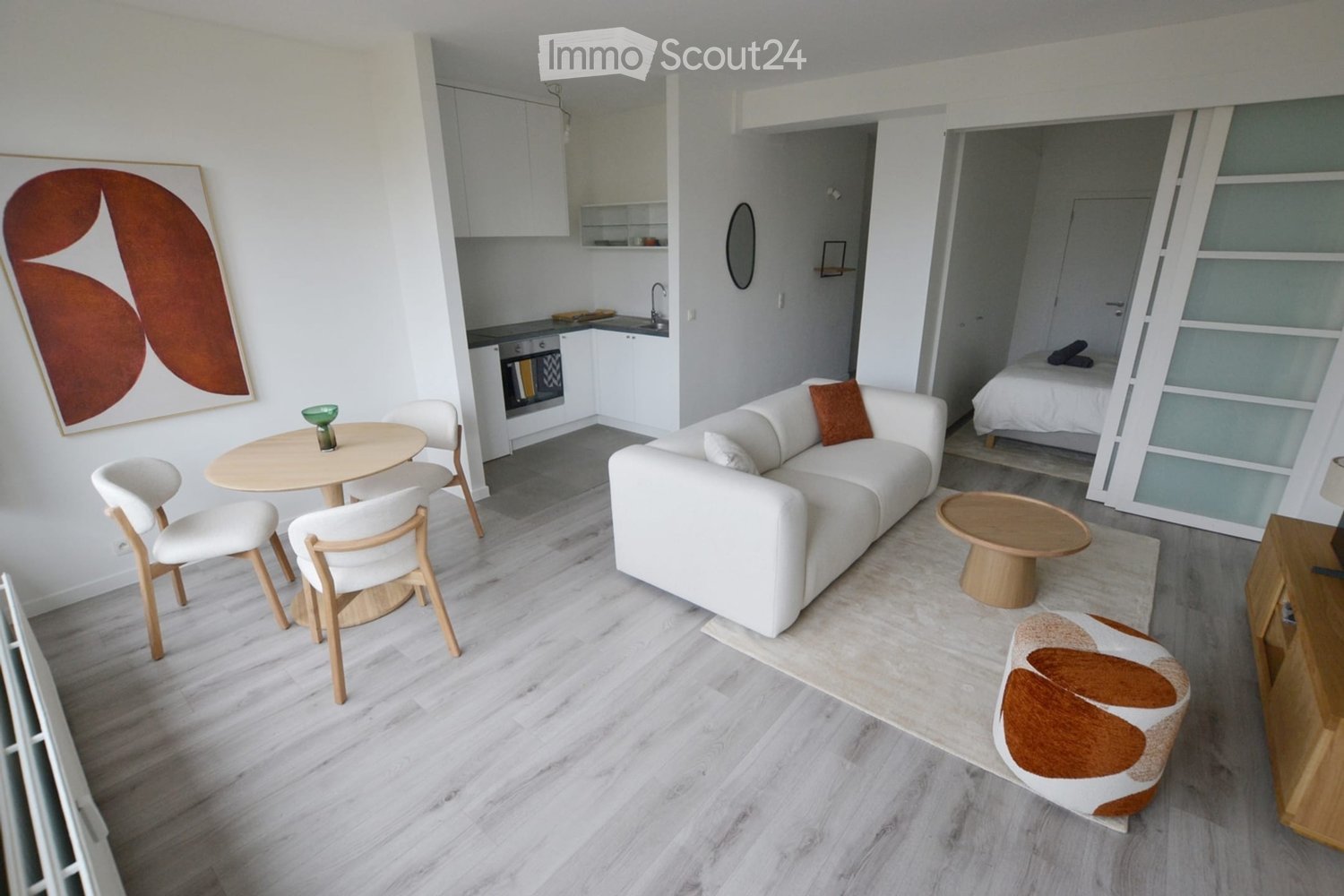 Modern, open-concept living space with a white sofa, coffee table, dining area with chairs and table, kitchen area with cabinets and oven, and a separate bedroom with a bed.