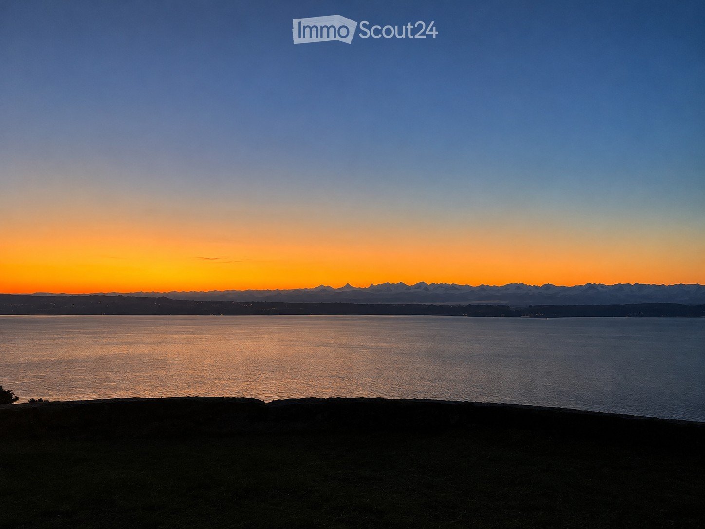 A serene view of a body of water at sunset, with distant mountains and a gradient sky transitioning from deep blue to warm orange hues.