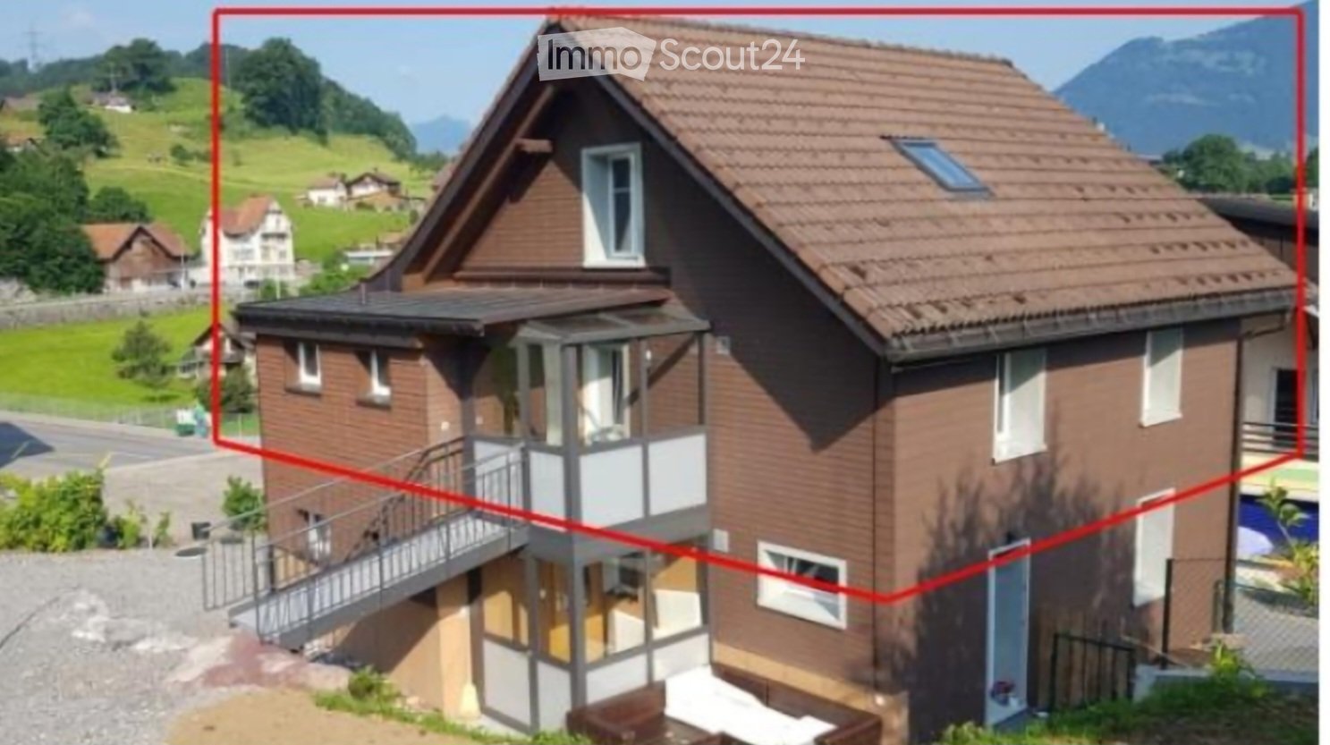 Two-story house with brown brick exterior, multiple windows, and a balcony on the first floor. The house features a sloped roof with solar panels and is situated in a scenic, green area with hills and trees in the background.