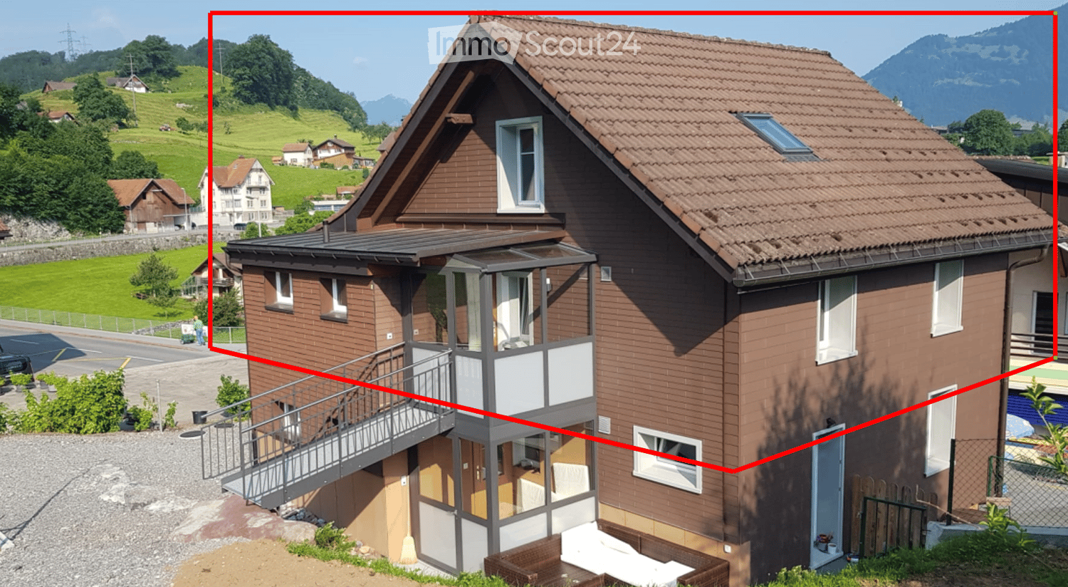 Two-story house with brown brick exterior, multiple windows, and a balcony on the first floor. The house is situated in a scenic, green landscape with hills and mountains in the background.