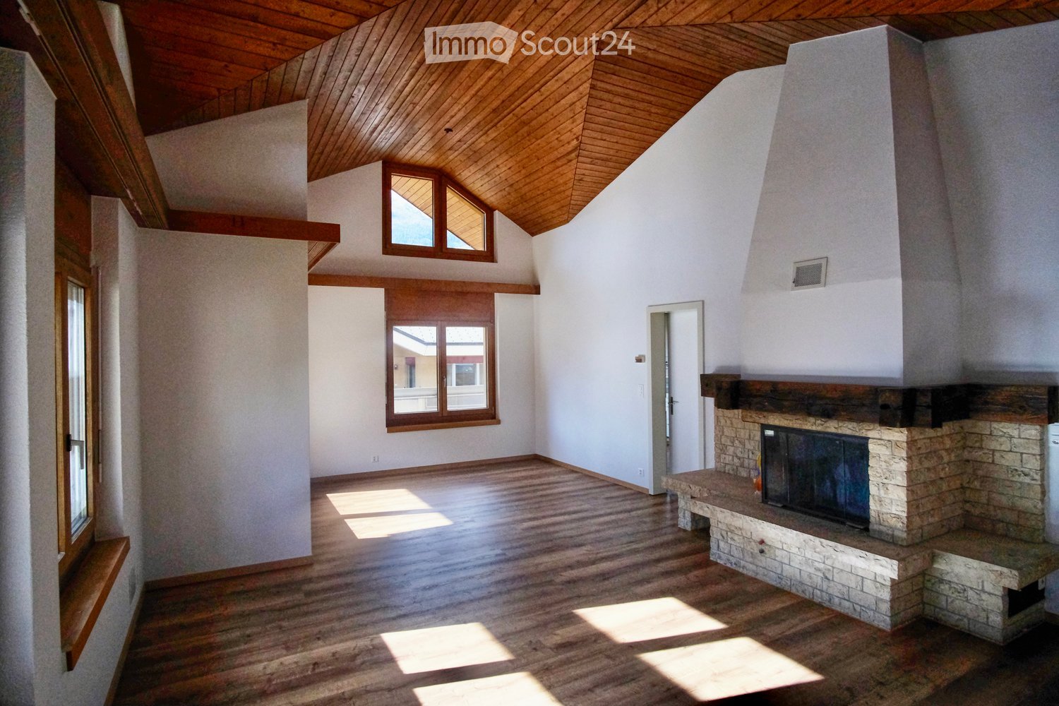 Empty room with wooden flooring, white walls, and a stone fireplace. The room features large windows and a wooden ceiling with exposed beams.