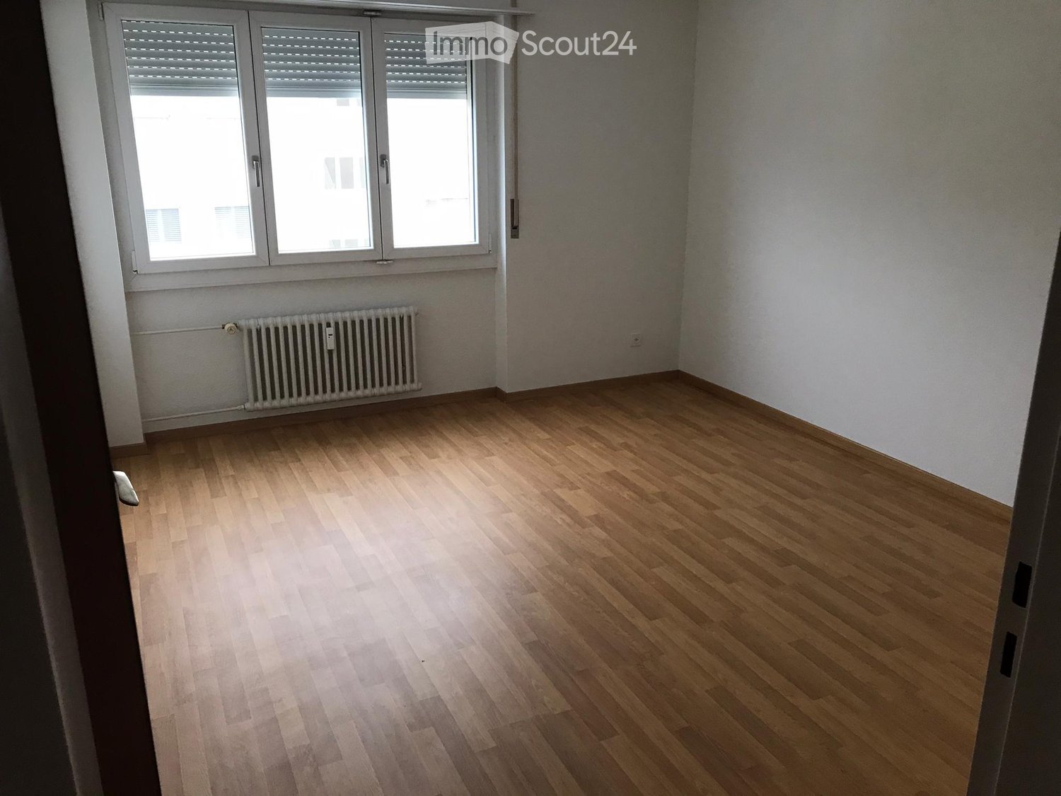 Empty room with light wooden flooring, white walls, and a large window with blinds. A radiator is visible below the window.