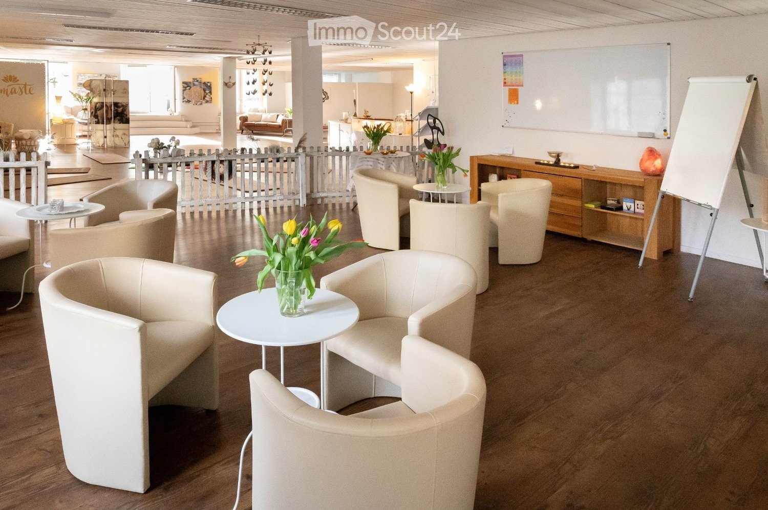 Modern, well-lit lounge area with several white chairs, small round tables, and vases with flowers. A large screen is visible on the right side.
