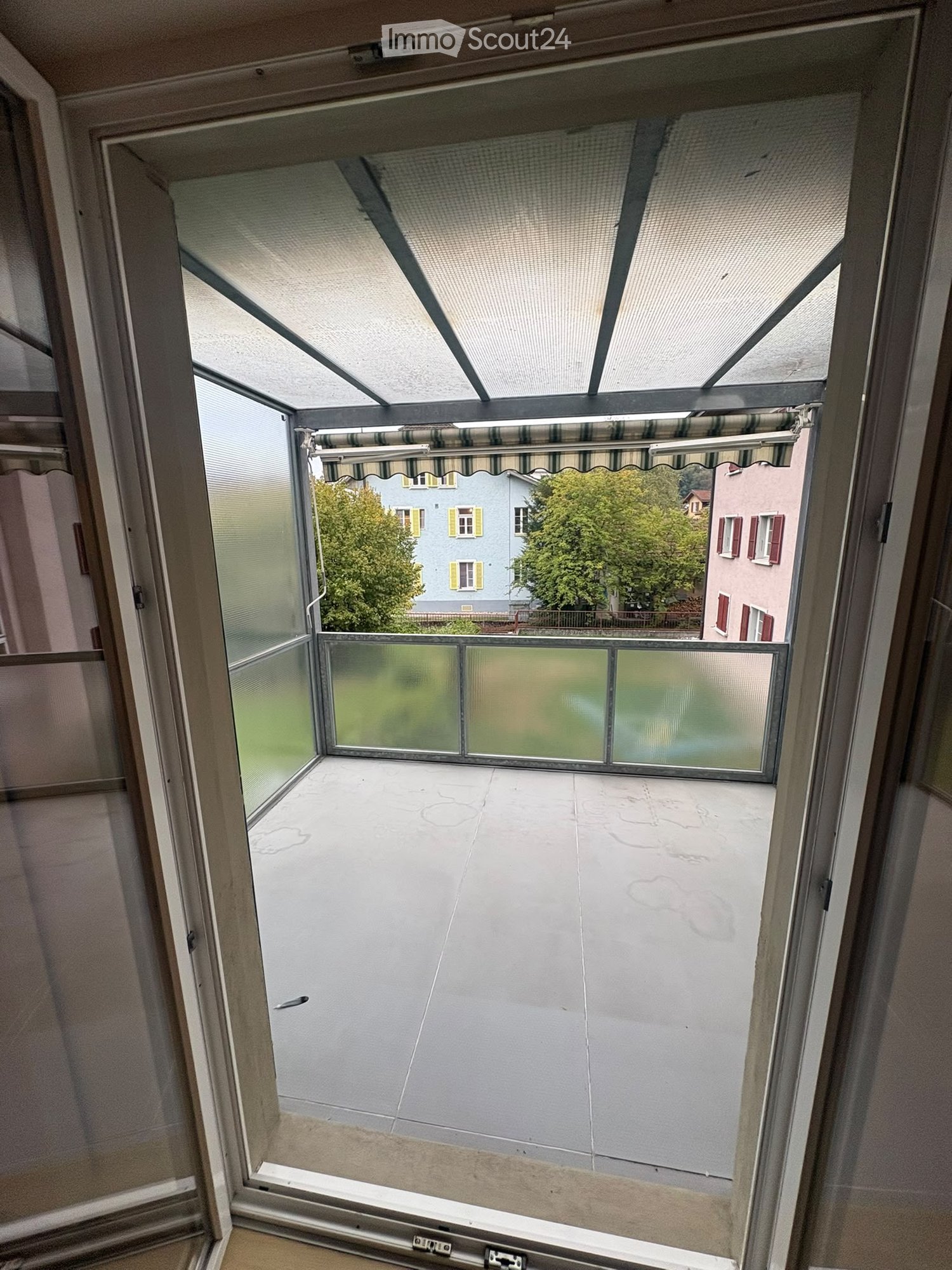 Balcony with glass enclosure, tiled floor, and retractable awning, overlooking a residential area with trees and houses.