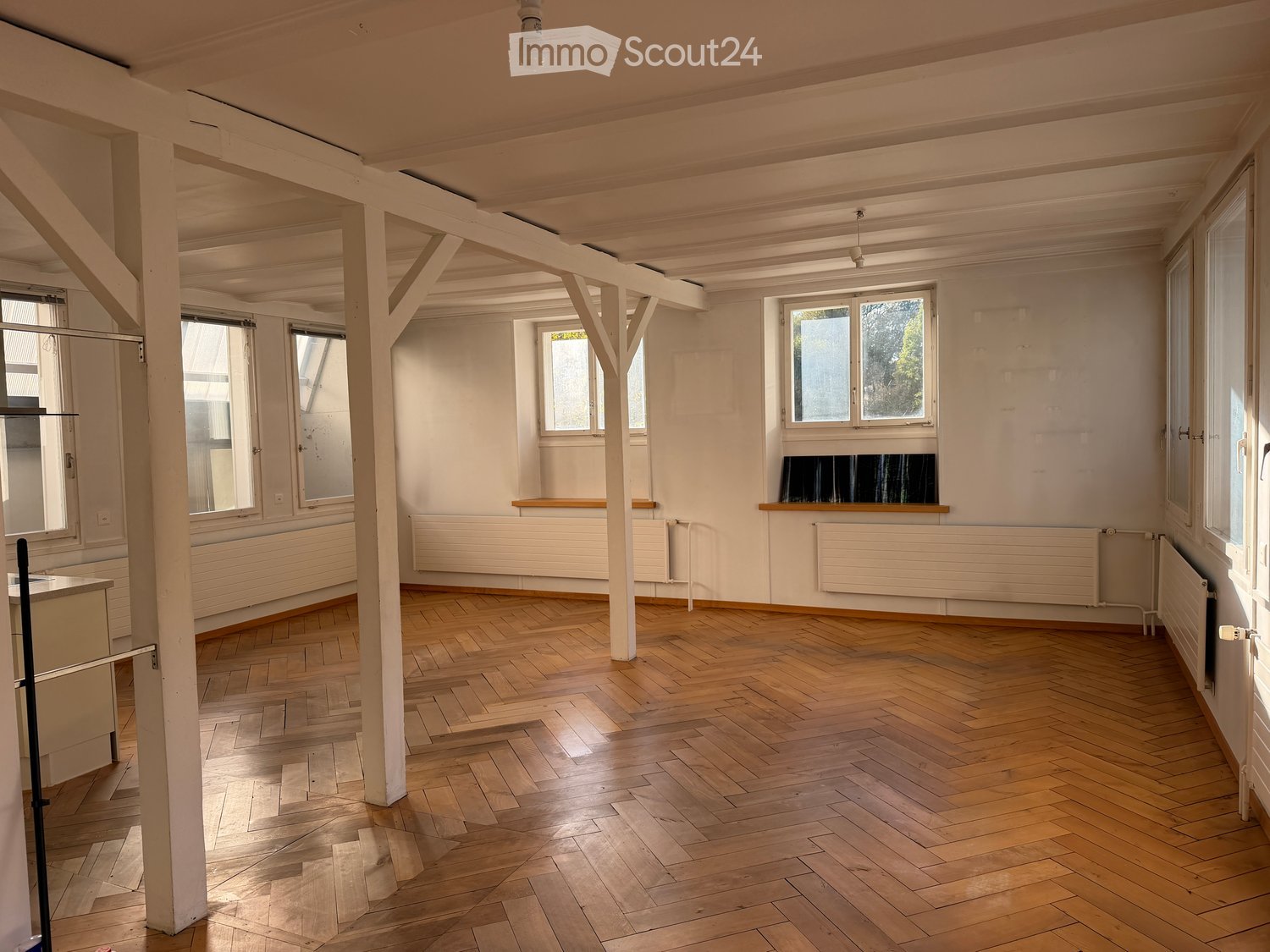 Empty room with wooden herringbone flooring, white walls, large windows, and exposed wooden beams. Radiators are visible along the walls.