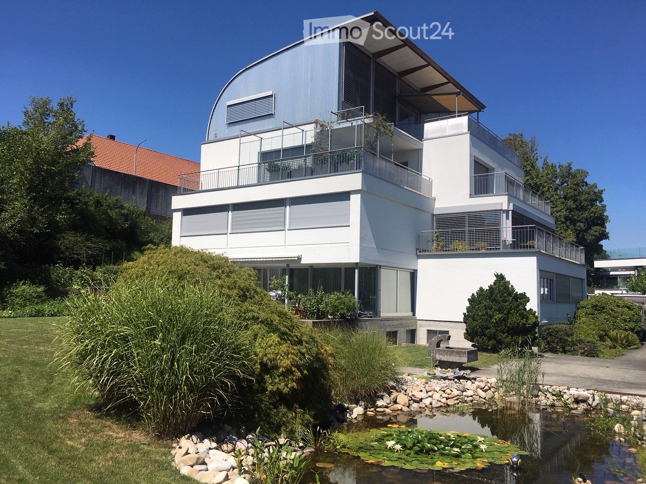 Modern two-story house with white exterior, curved roof, large windows, and balconies. The surrounding area includes a pond with lily pads, a stone bench, and landscaped greenery.