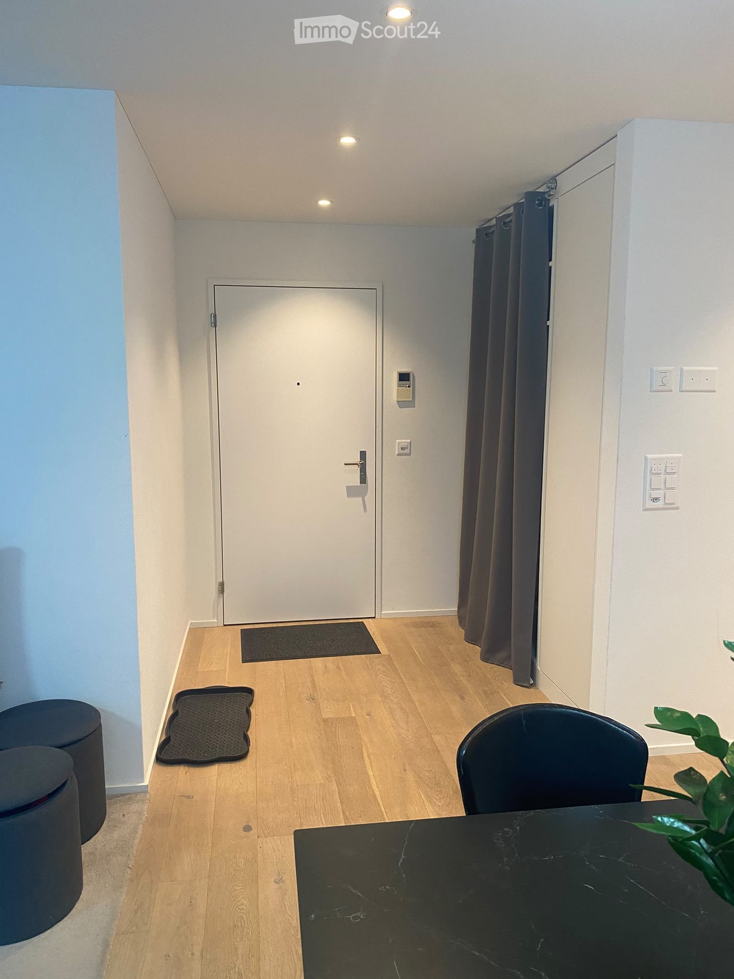 A small, modern hallway with wooden flooring, a black table, a chair, two stools, a door, and a curtain.