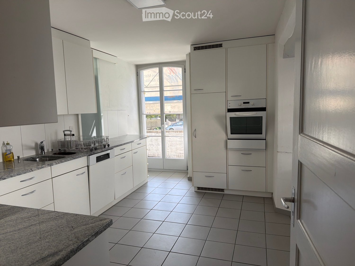 Modern kitchen with white cabinets, granite countertop, stainless steel sink, dishwasher, oven, refrigerator, and a bottle on the counter.