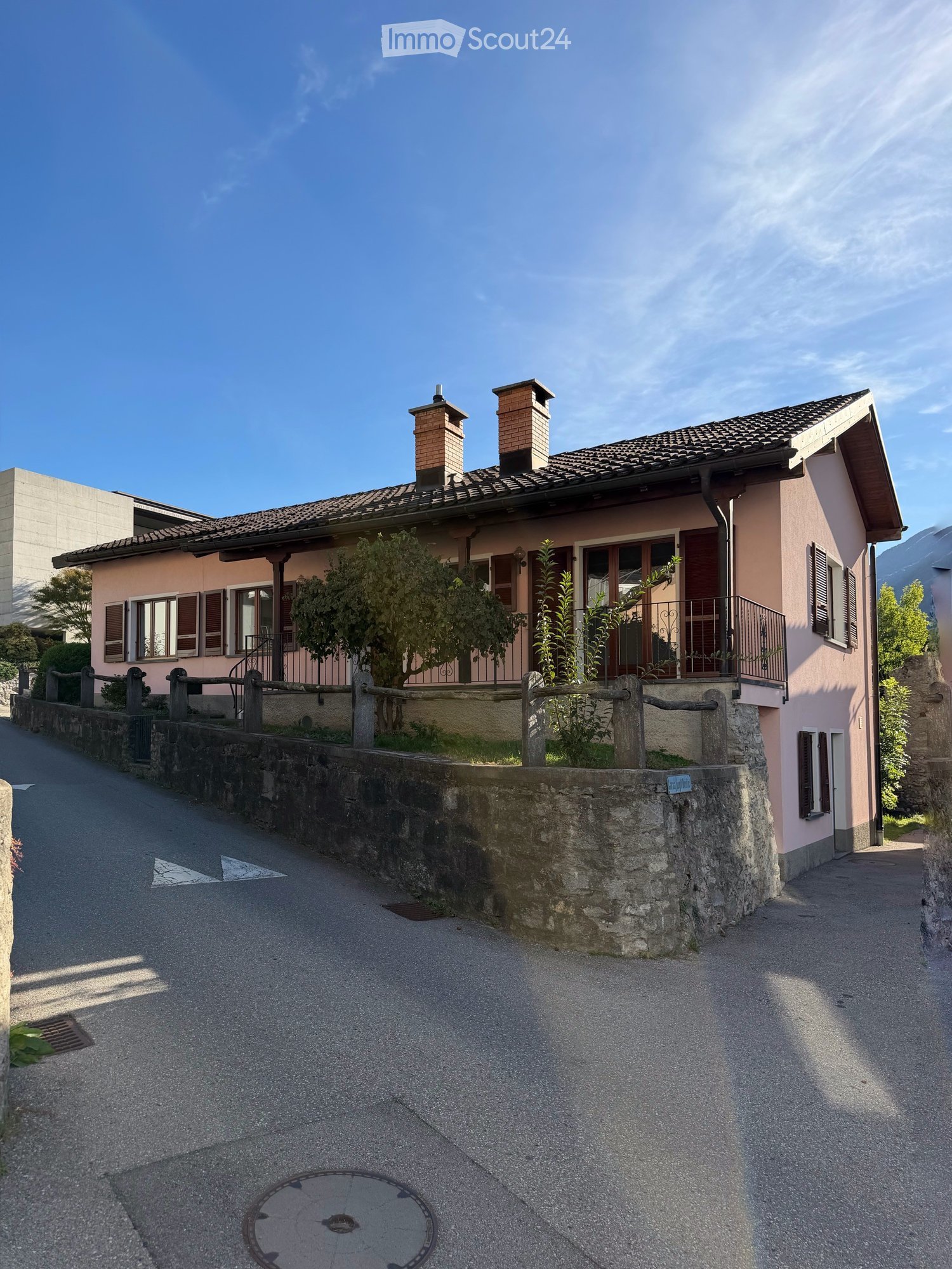 Two-story house with pink exterior, tiled roof, chimneys, and wooden shutters. Features a stone wall and small garden area with plants and a wooden fence.