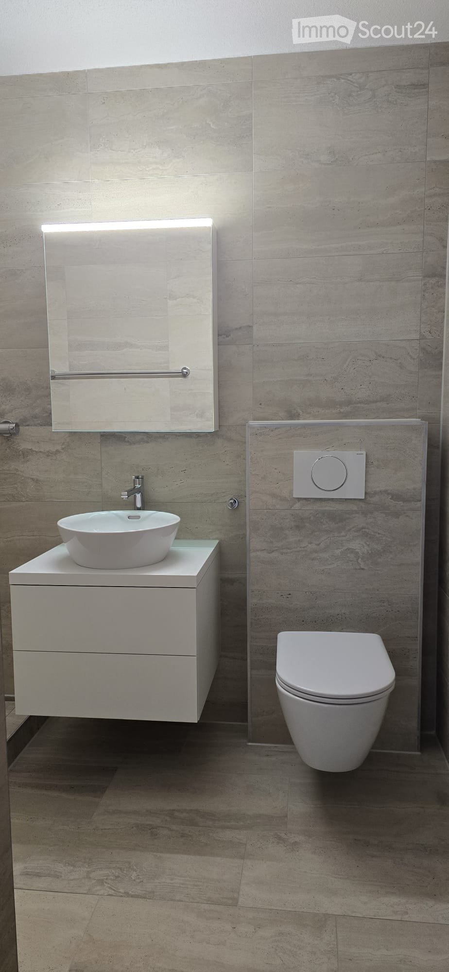 Modern bathroom with a white sink, toilet, and mirrored cabinet, featuring light grey tiled walls and a wooden floor.