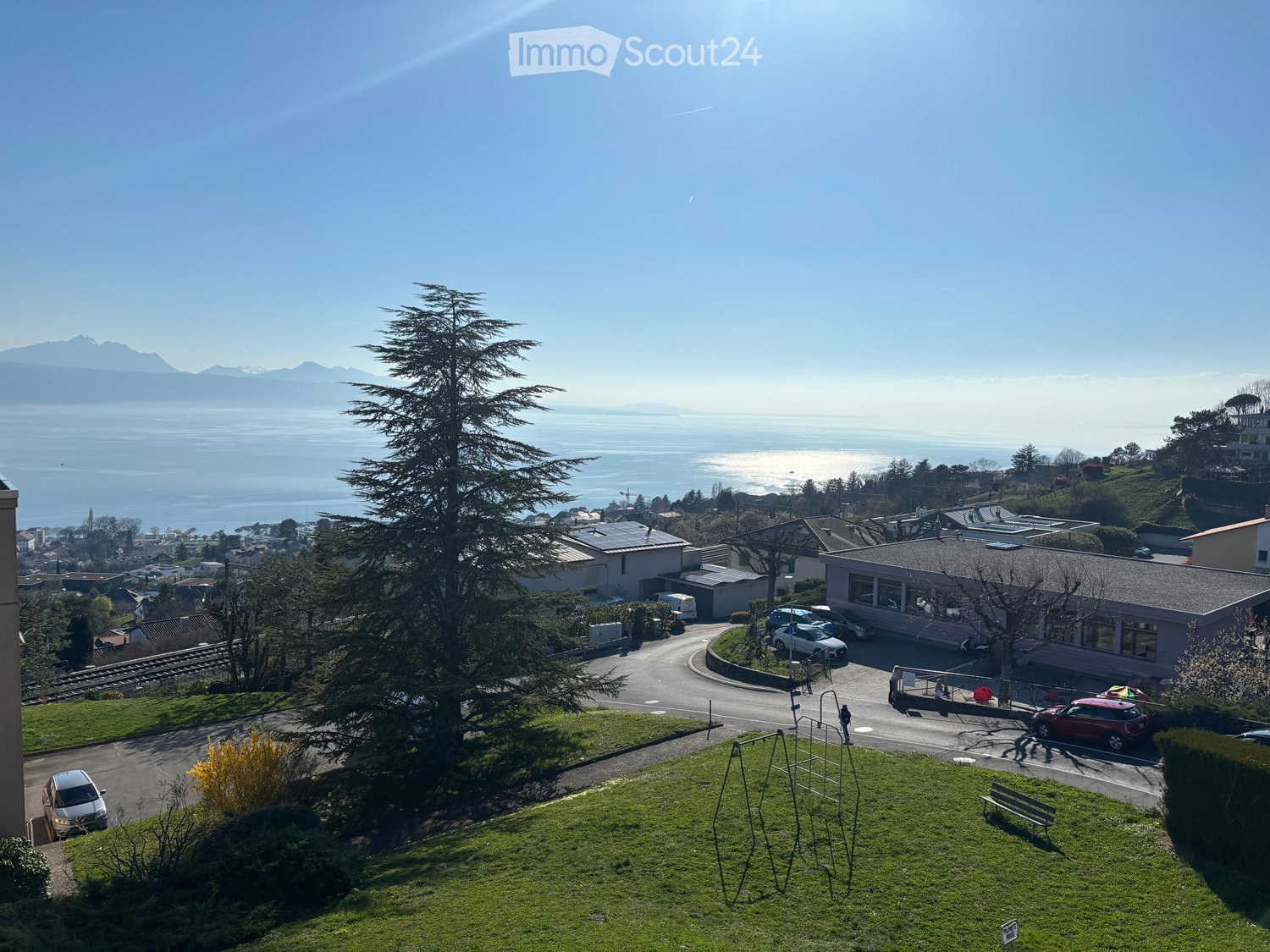 A scenic view of a residential area with a large tree in the foreground, a playground, parked cars, and a distant lake and mountains.
