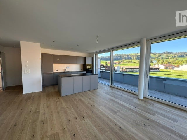 Modern kitchen with light grey cabinets, a central island, and large windows offering a scenic view of the countryside.