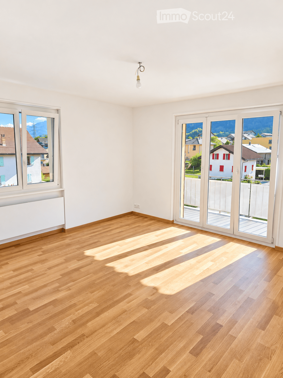 Empty room with light wooden flooring, white walls, large windows, and sliding glass doors leading to a balcony.