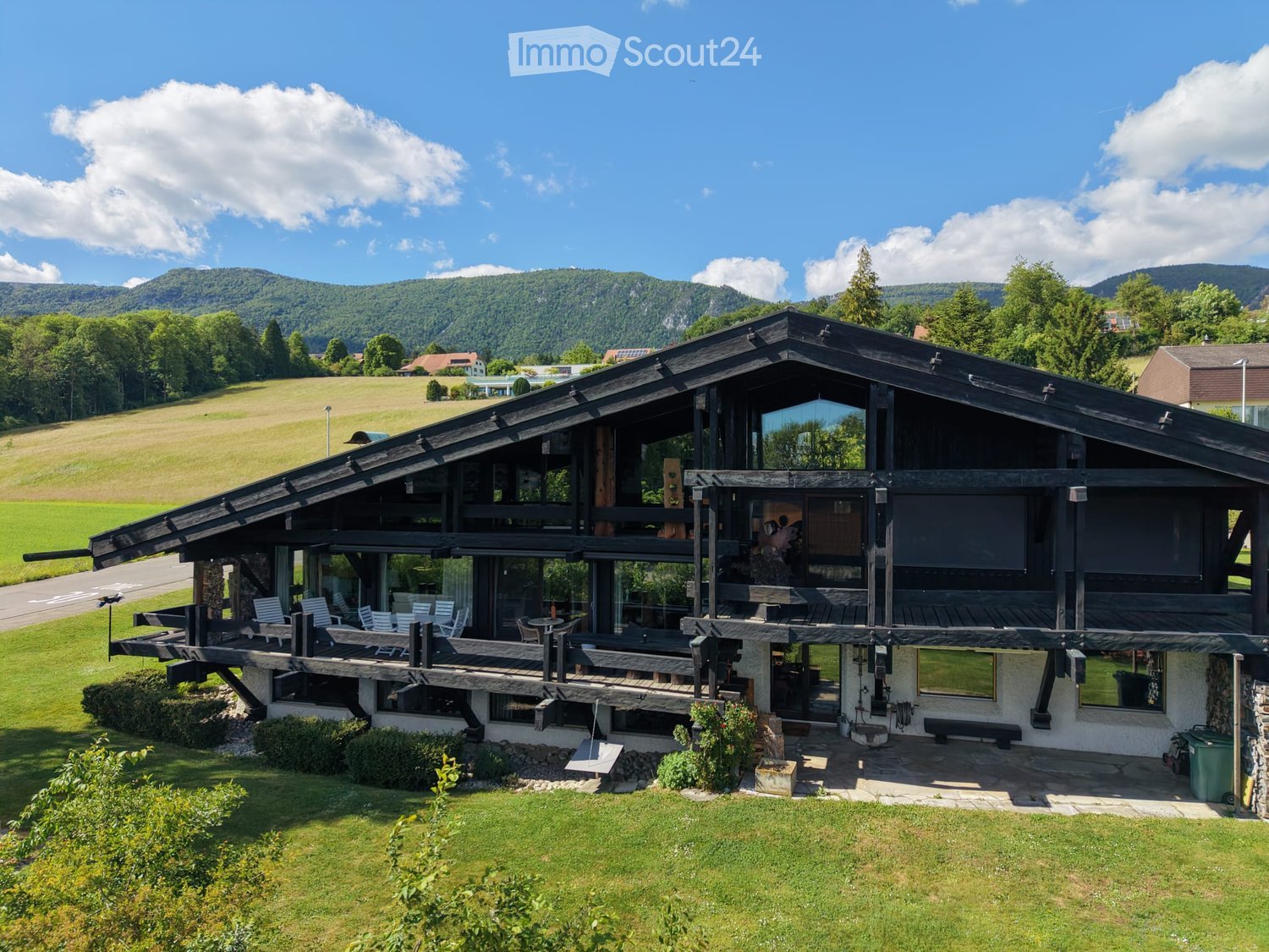 Two-story wooden house with large windows and a spacious wooden deck, surrounded by a well-maintained lawn and scenic mountain backdrop.