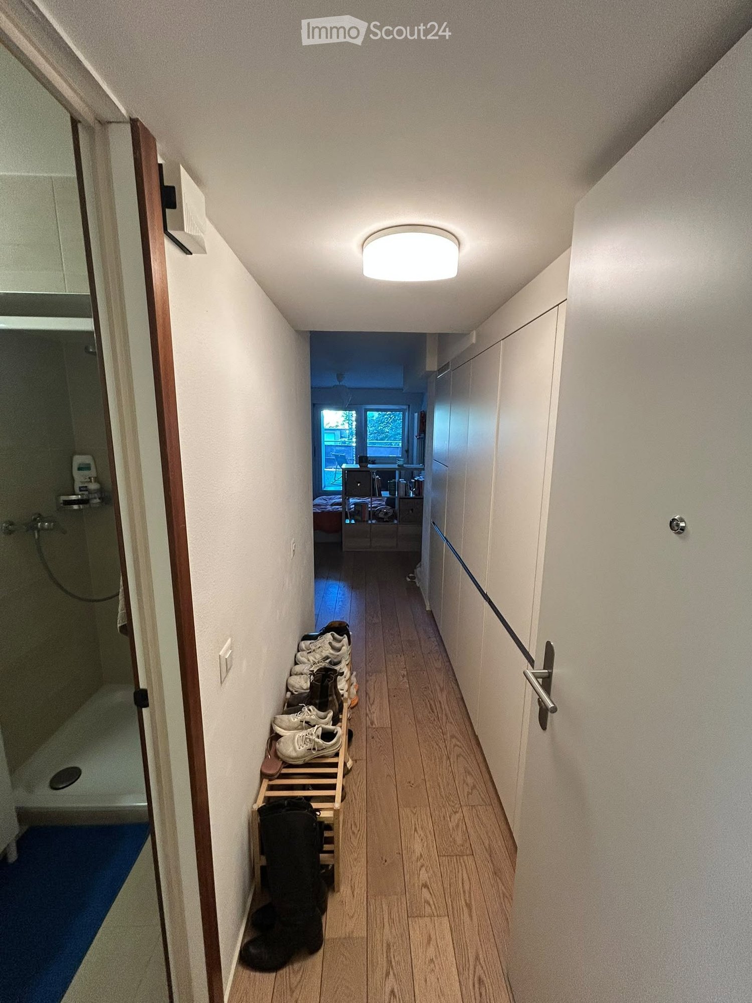 A narrow hallway with wooden flooring, white walls, and a ceiling light. Shoes are placed on a small wooden shelf on the left side, and a partially visible bathroom is on the left. The hallway leads to a room with a bed and a window at the end.