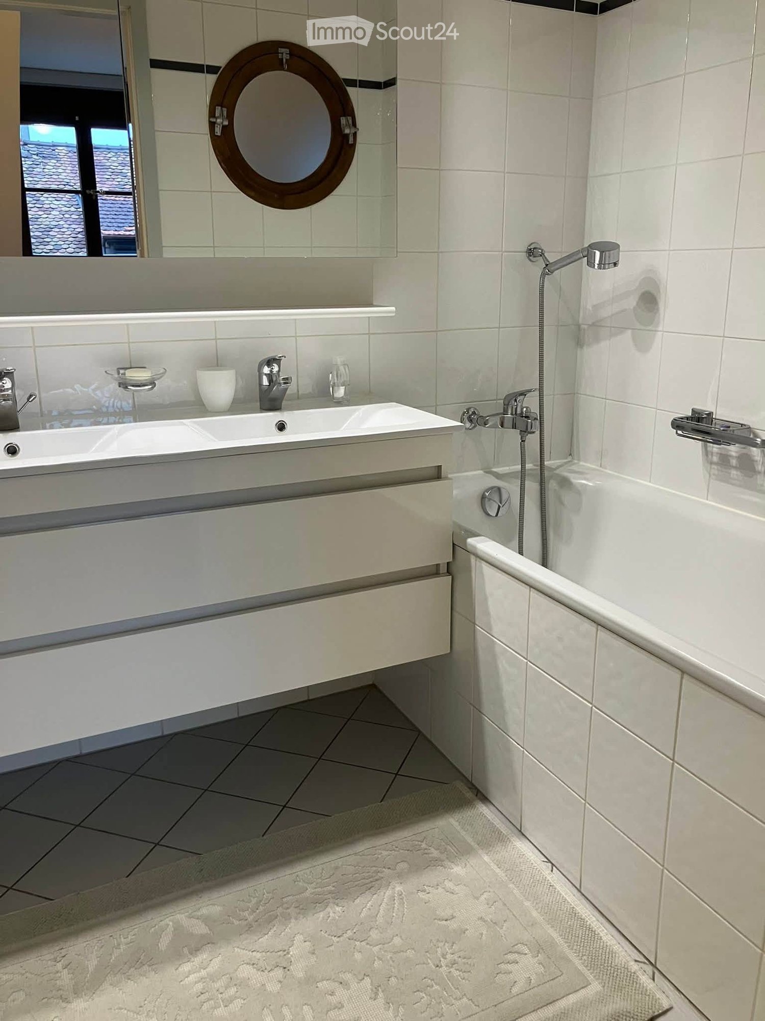 Modern bathroom with a double sink, bathtub, and a round mirror with a wooden frame. The sink has a sleek design with a built-in drawer, and the bathtub features a handheld showerhead.