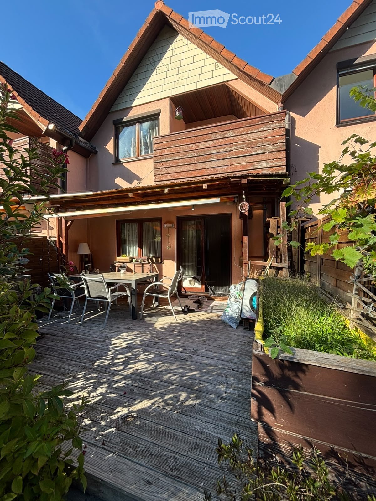 Wooden deck area with a table and chairs, surrounded by greenery and a wooden fence, with a house featuring a balcony in the background.