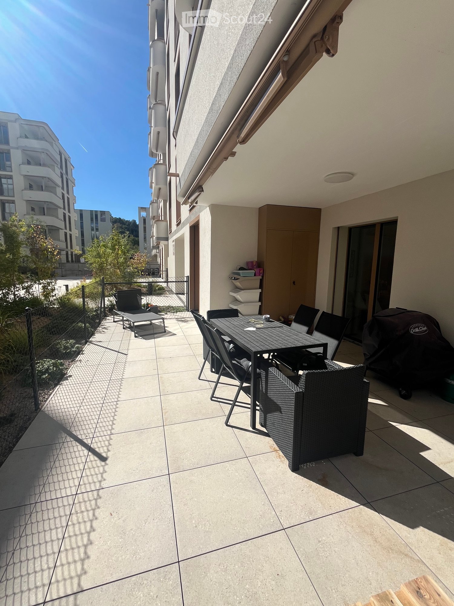 Balcony with tiled floor, outdoor furniture including a table, chairs, and an umbrella, with a view of neighboring buildings and greenery.