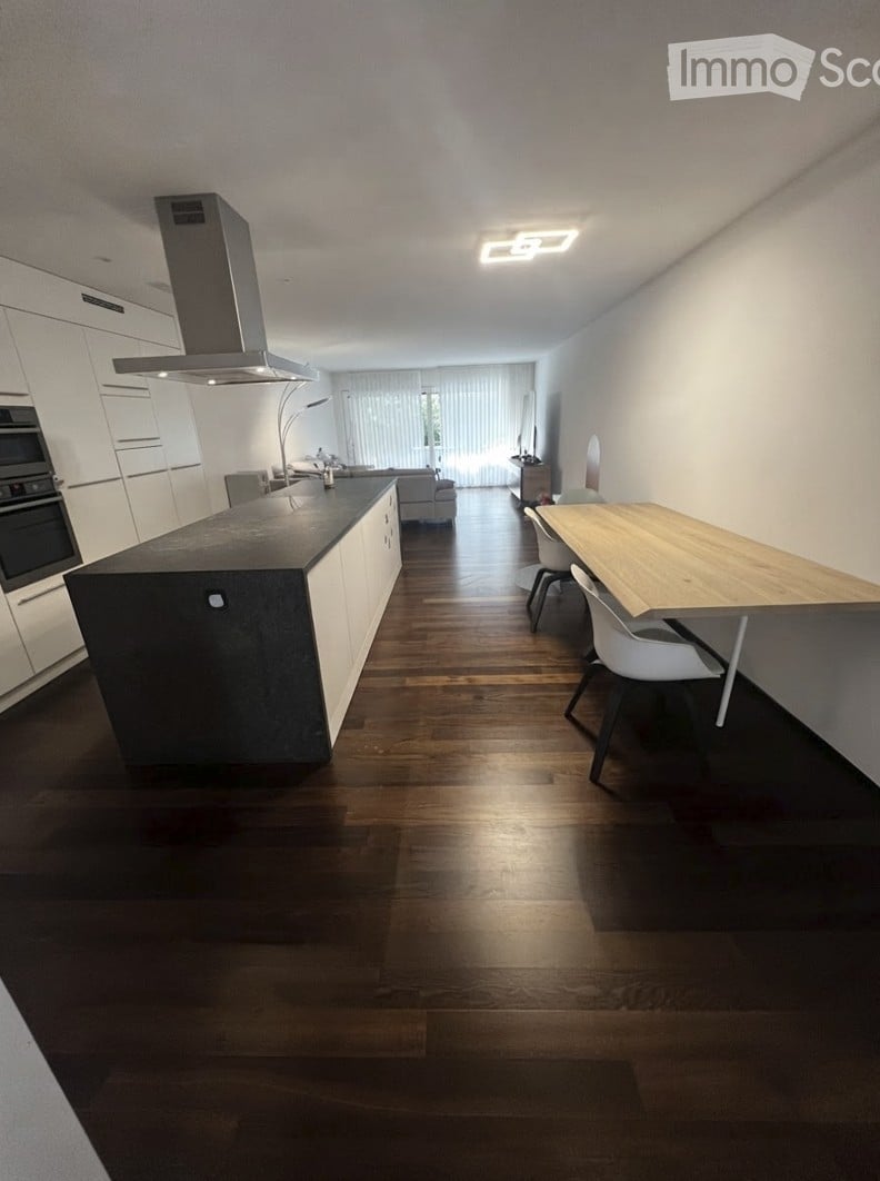 Modern kitchen with a dark countertop island, white cabinets, built-in oven, range hood, and a dining area with a wooden table and chairs. The space features hardwood flooring and large windows with blinds.