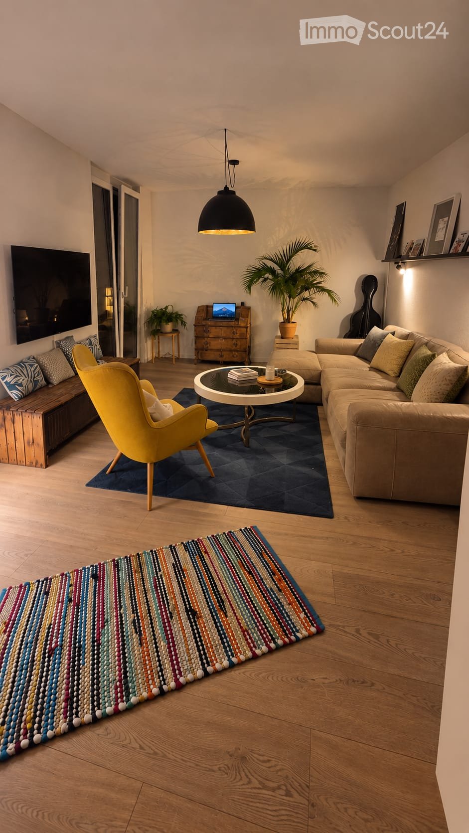 Modern living room with wooden flooring, a large sectional sofa, a yellow armchair, a round coffee table, a laptop on a small table, a potted plant, a guitar, and a colorful beaded runner rug.