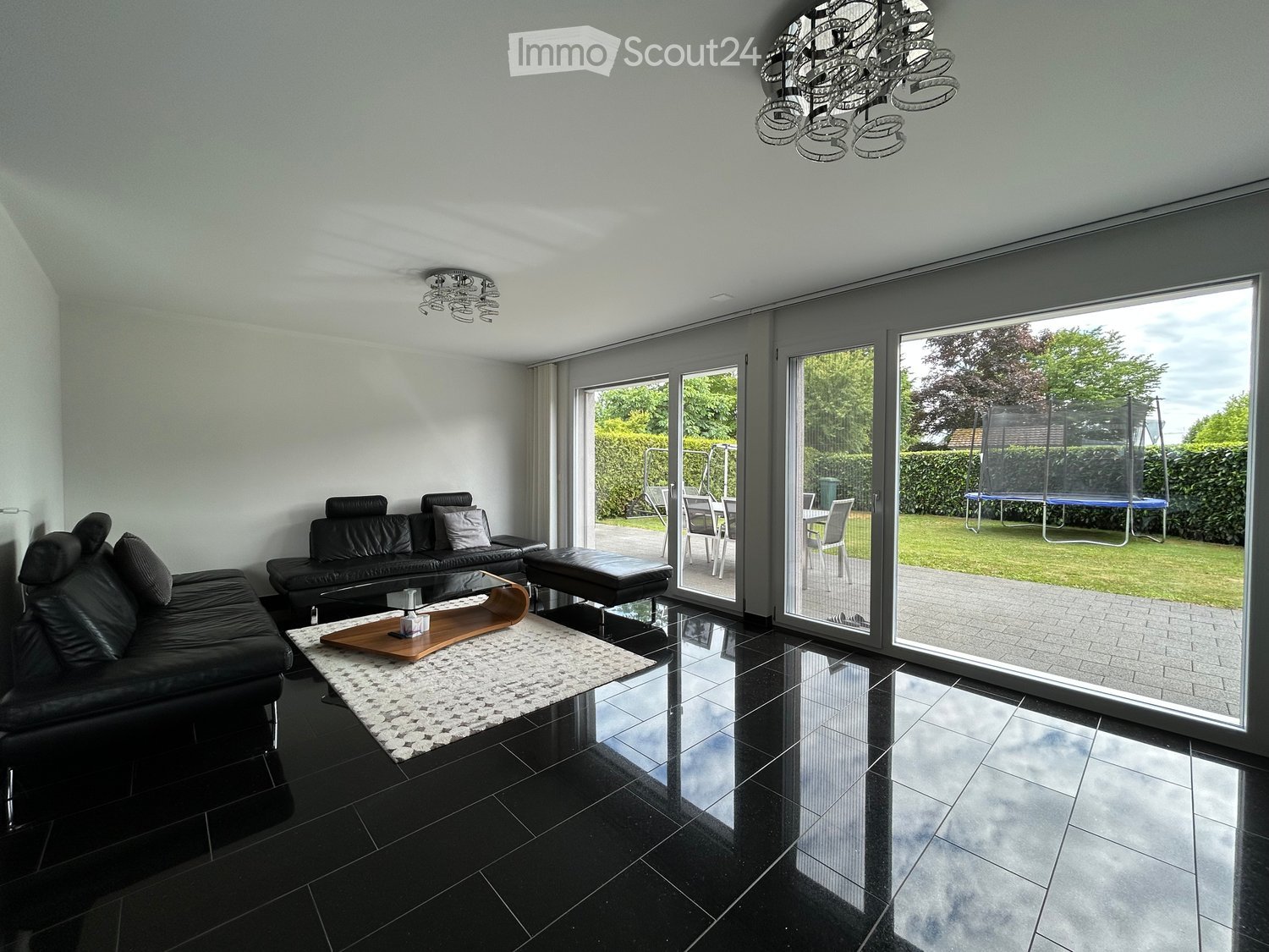 Modern living room with black tiled flooring, large sliding glass doors leading to a patio, a black leather sofa, a wooden coffee table, and an ottoman. The room features contemporary lighting fixtures and a view of a well-maintained garden with outdoor f
