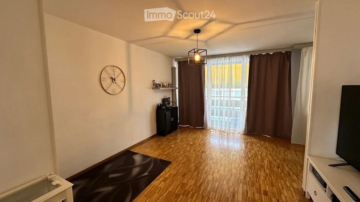 A modern living room with wooden flooring, a large window with curtains, a clock on the wall, a shelf with items, a small cabinet, a table with a chair, and a television on a stand.