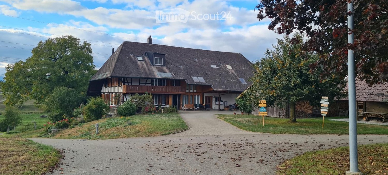 Two-story house with a sloped roof, wooden balcony, and solar panels on the roof. The house is surrounded by a well-maintained lawn, trees, and a gravel driveway.