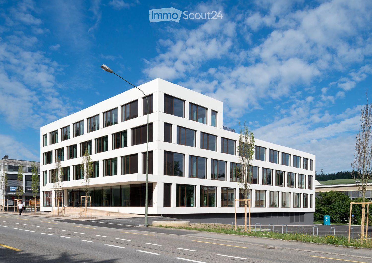 Modern, white, multi-story building with large windows and a clean, contemporary design. The building is situated on a street with a sidewalk and streetlight nearby.