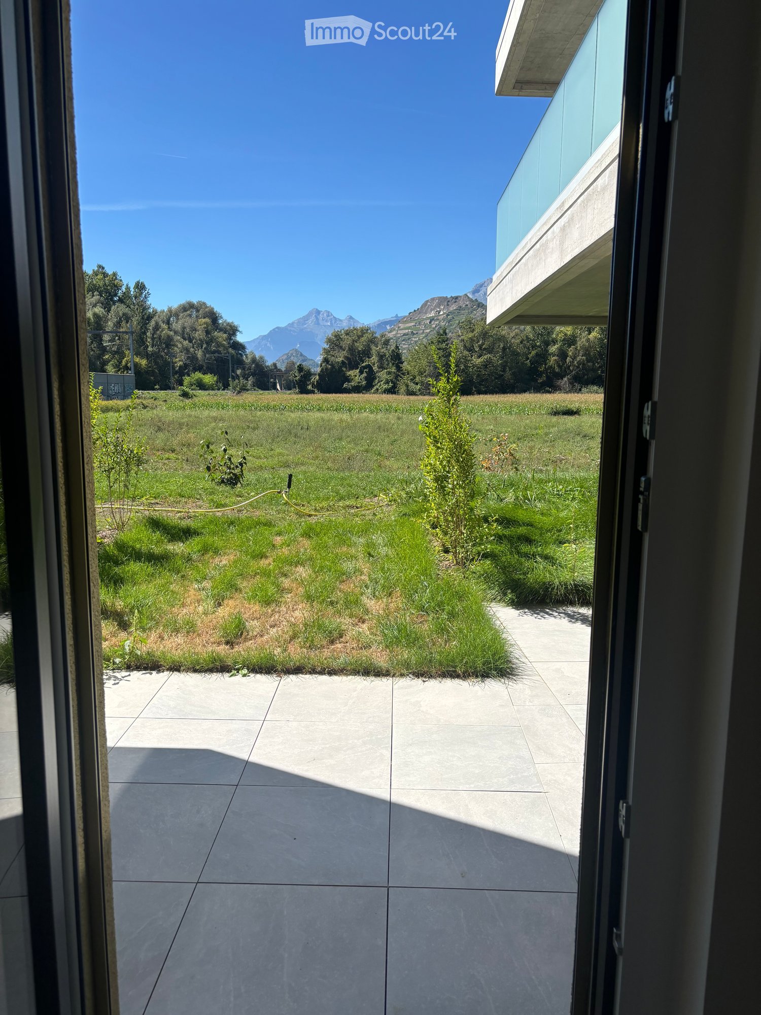View from inside a house onto a well-maintained garden with a lawn, small plants, and a modern building with a glass facade in the background, under a clear blue sky with distant mountains.