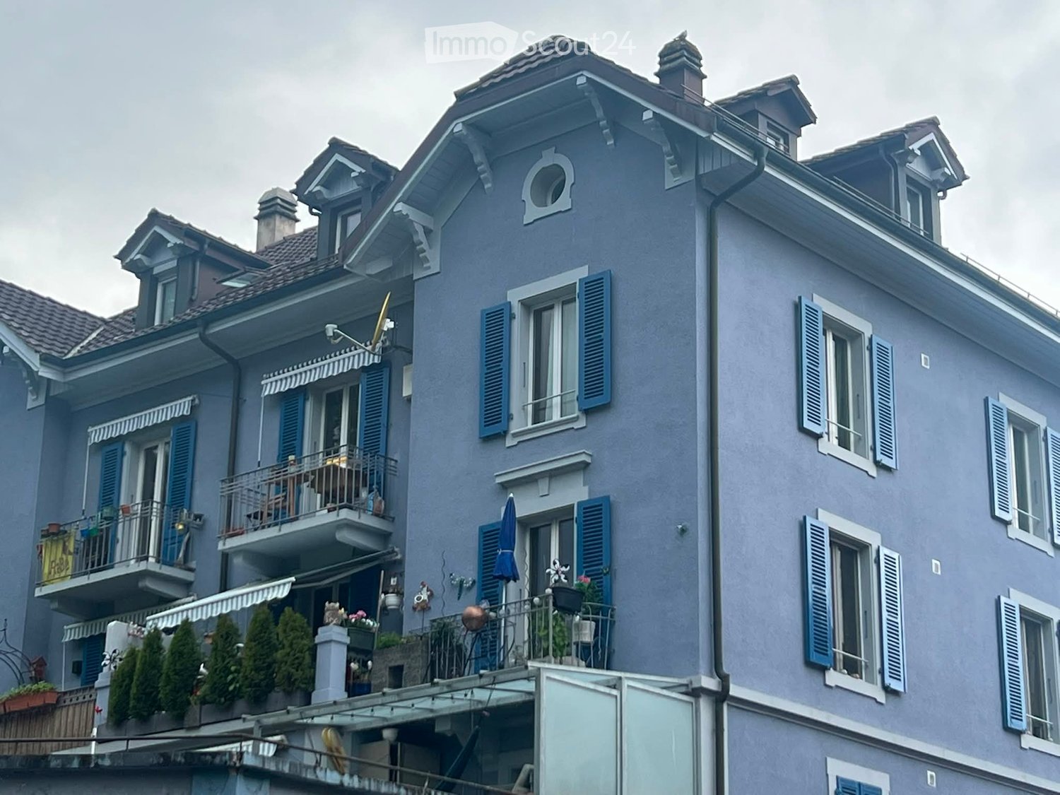 A multi-story residential building with blue shutters, balconies, and decorative elements. The facade features multiple windows, a chimney, and a roof with dormer windows.