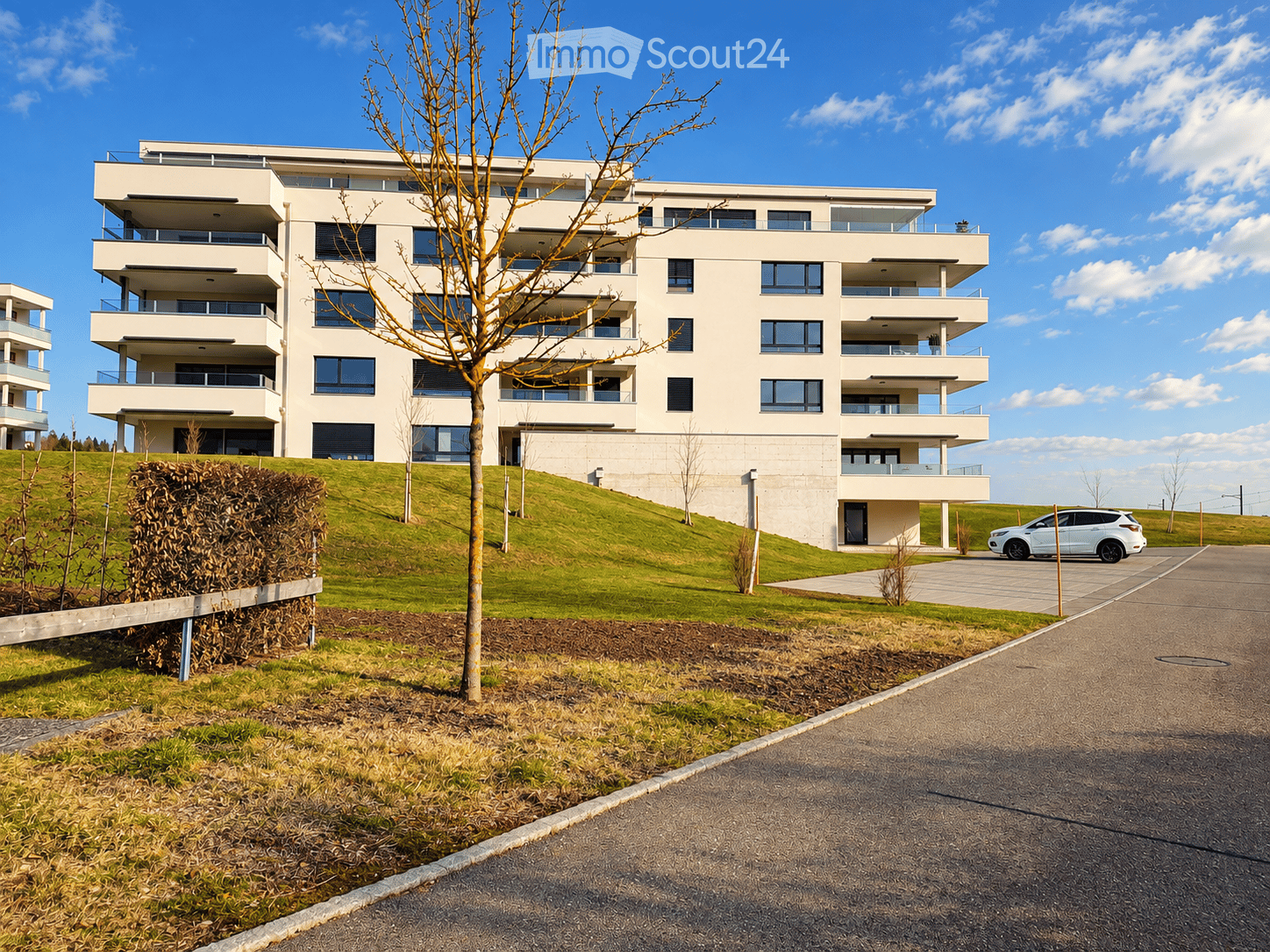 Modern multi-story residential building with balconies and large windows, surrounded by a landscaped area with a tree and a parked car.