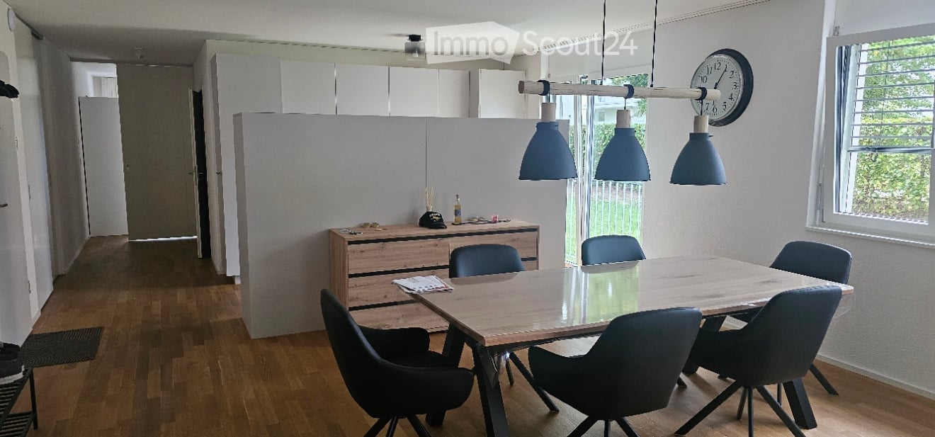 A modern, well-lit room with a wooden dining table surrounded by chairs, a wooden cabinet with various items, and a clock on the wall. The room features wooden flooring and large windows allowing natural light to fill the space.