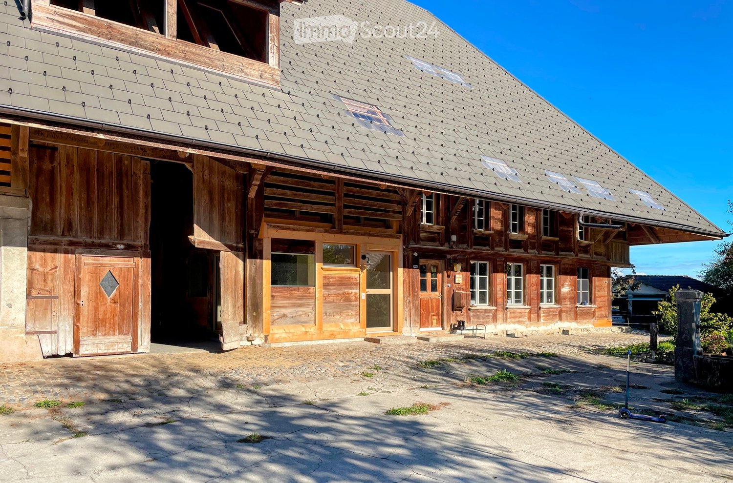 A rustic wooden building with a steep, grey tiled roof and multiple windows, featuring large wooden doors and a stone-paved courtyard.