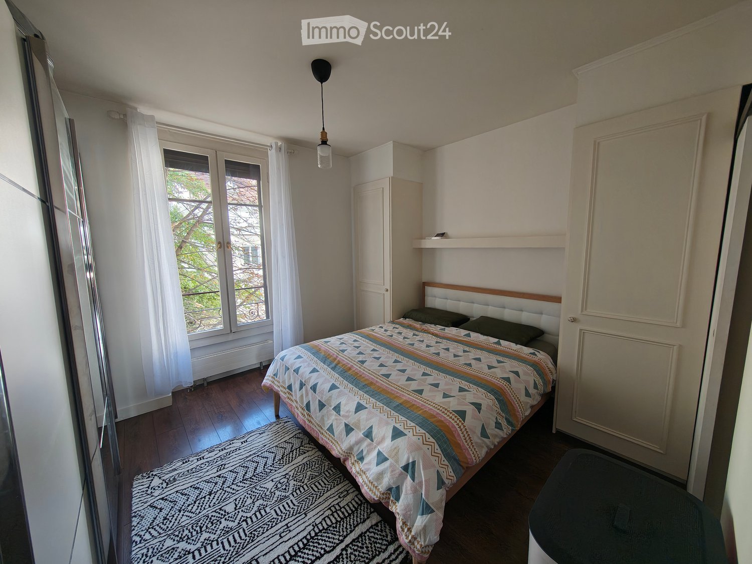 Cozy bedroom with a wooden bed, colorful bedspread, nightstand, wardrobe, and a large window with white curtains.