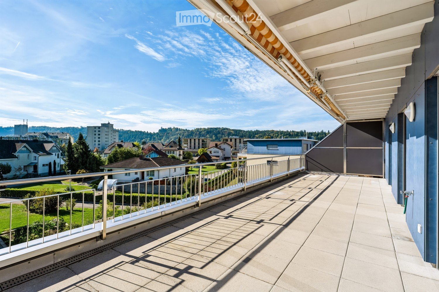 Balcony with tiled floor, metal railing, and retractable awning, overlooking a residential area with houses and greenery.