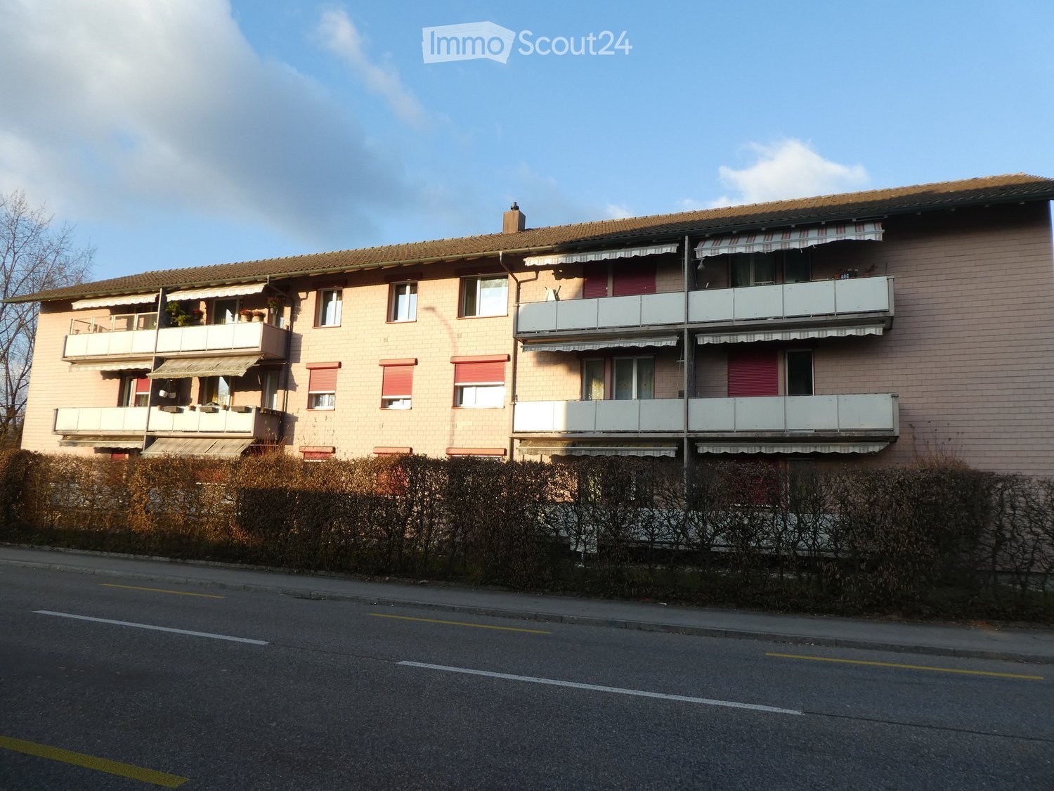 Multi-story residential building with balconies, awnings, and a tiled roof, surrounded by bushes and a road.