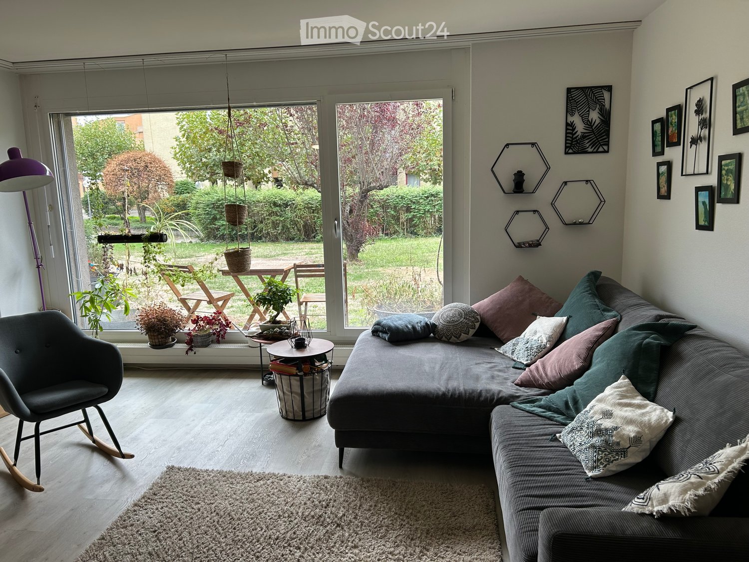 Modern living room with large windows overlooking a garden, featuring a grey sectional sofa with colorful pillows, a rocking chair, a coffee table, a floor lamp, and several potted plants on the windowsill.