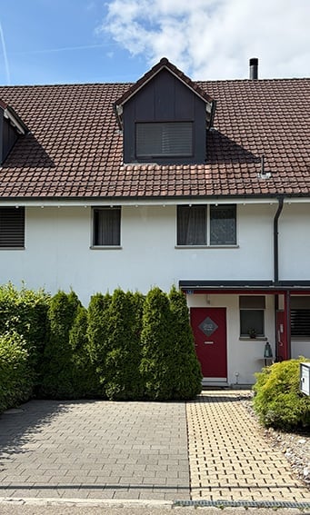 Two-story house with a red-tiled roof, white exterior walls, and a central dormer window. Features a red front door, multiple windows, and a neatly trimmed hedge along the front yard.