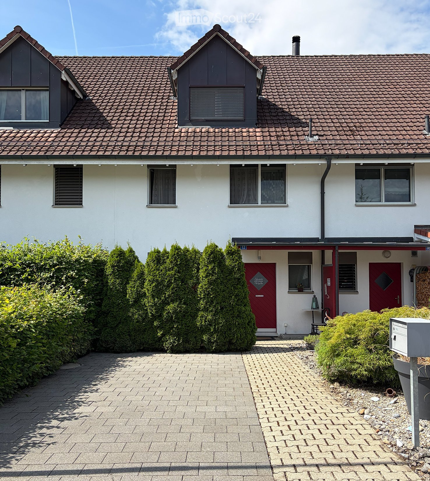 Two-story house with red-tiled roof, white walls, multiple windows, and a red front door. The house is surrounded by neatly trimmed hedges and a paved driveway.