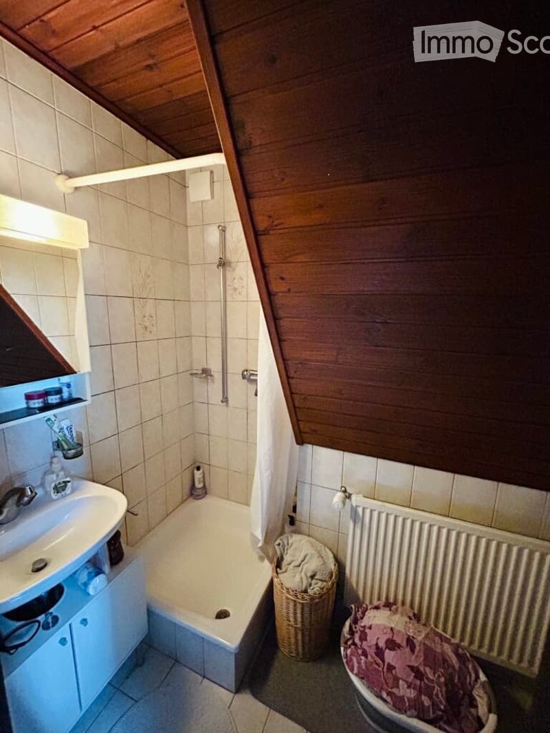 Bathroom with a wooden slanted ceiling, tiled walls, a white sink with a mirror, a bathtub, a radiator, and a wicker basket on the floor.
