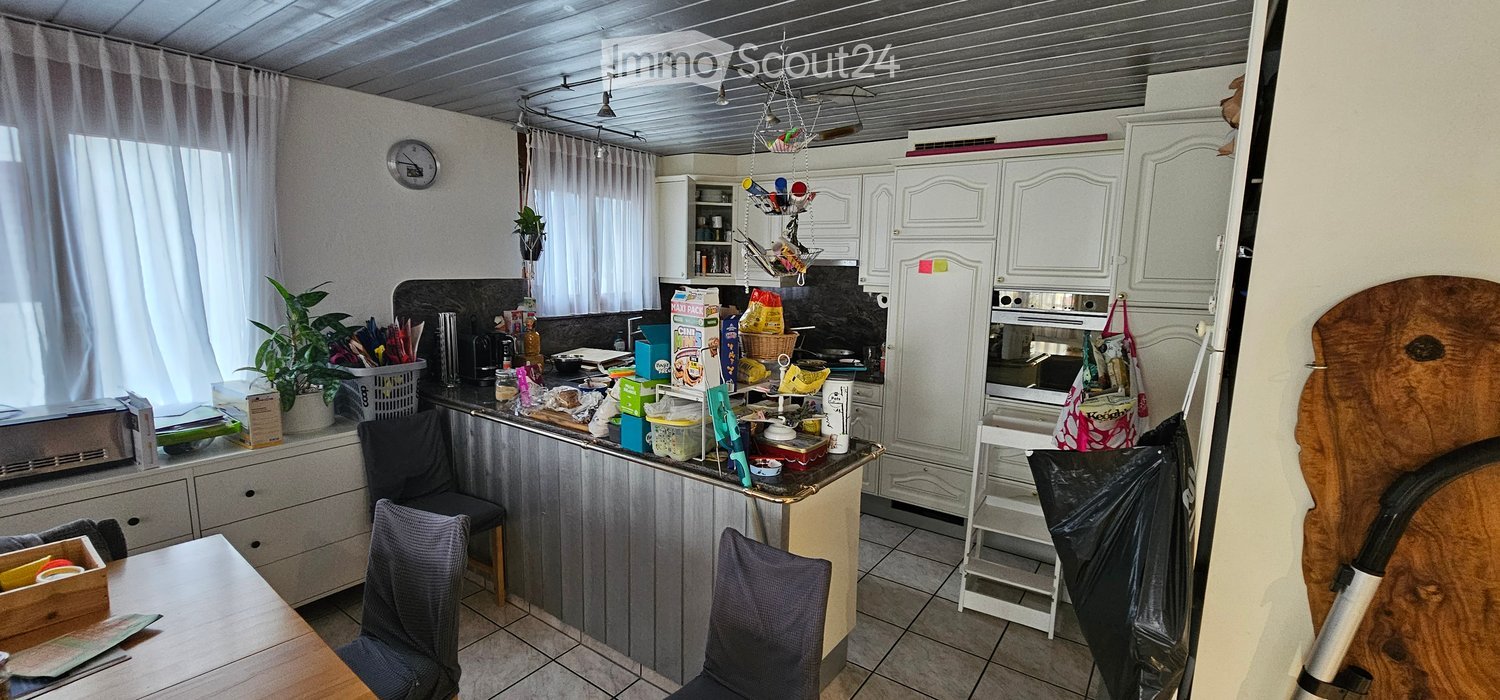 The kitchen features white cabinets, a refrigerator, an oven, and a microwave. There are chairs, a table, and a clock on the wall. The counter is cluttered with various items, including a basket, a plant, and some food packages.