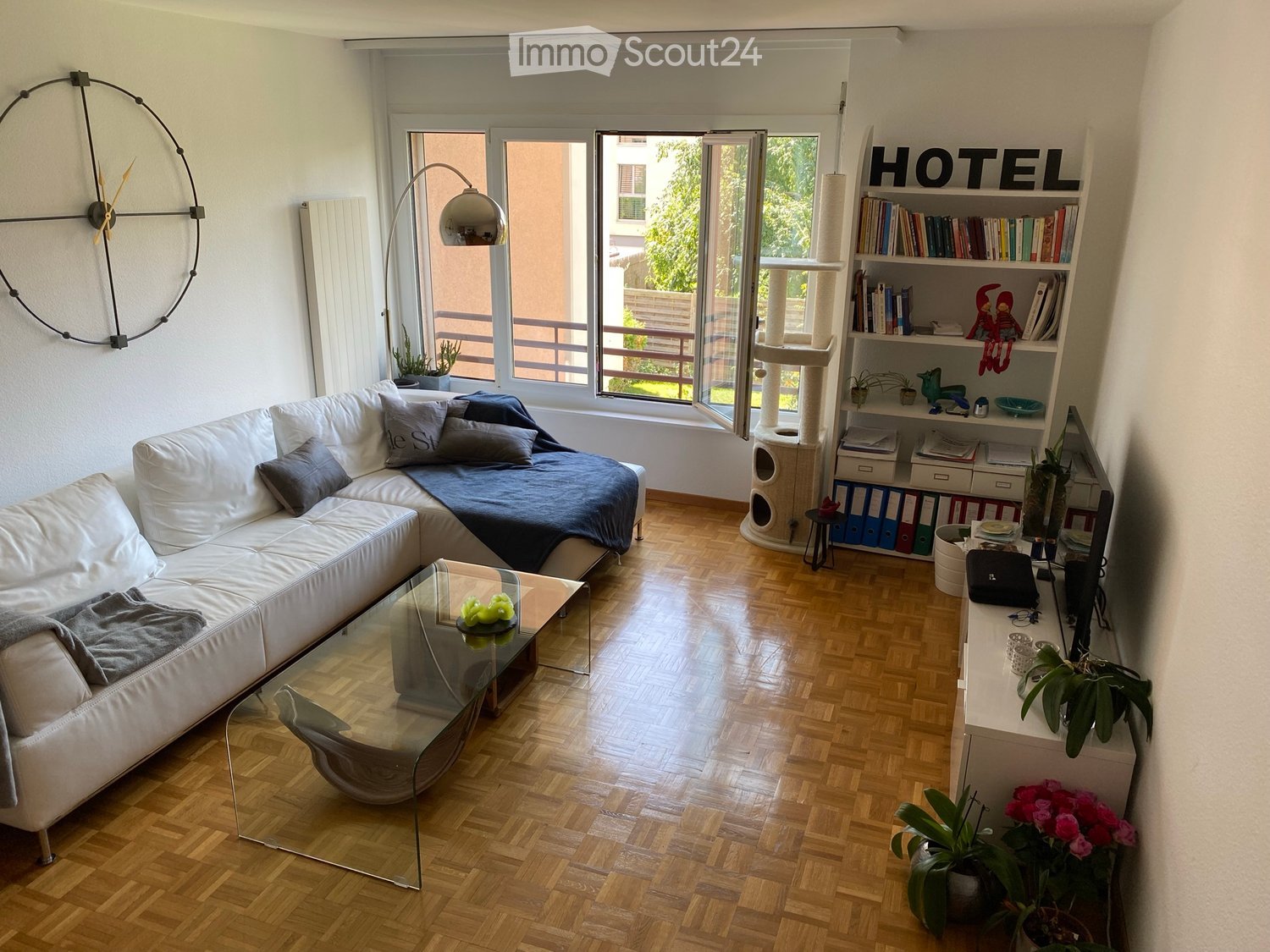 Modern living room with a white leather sofa, glass coffee table, bookshelf, cat tree, and large windows leading to a balcony.