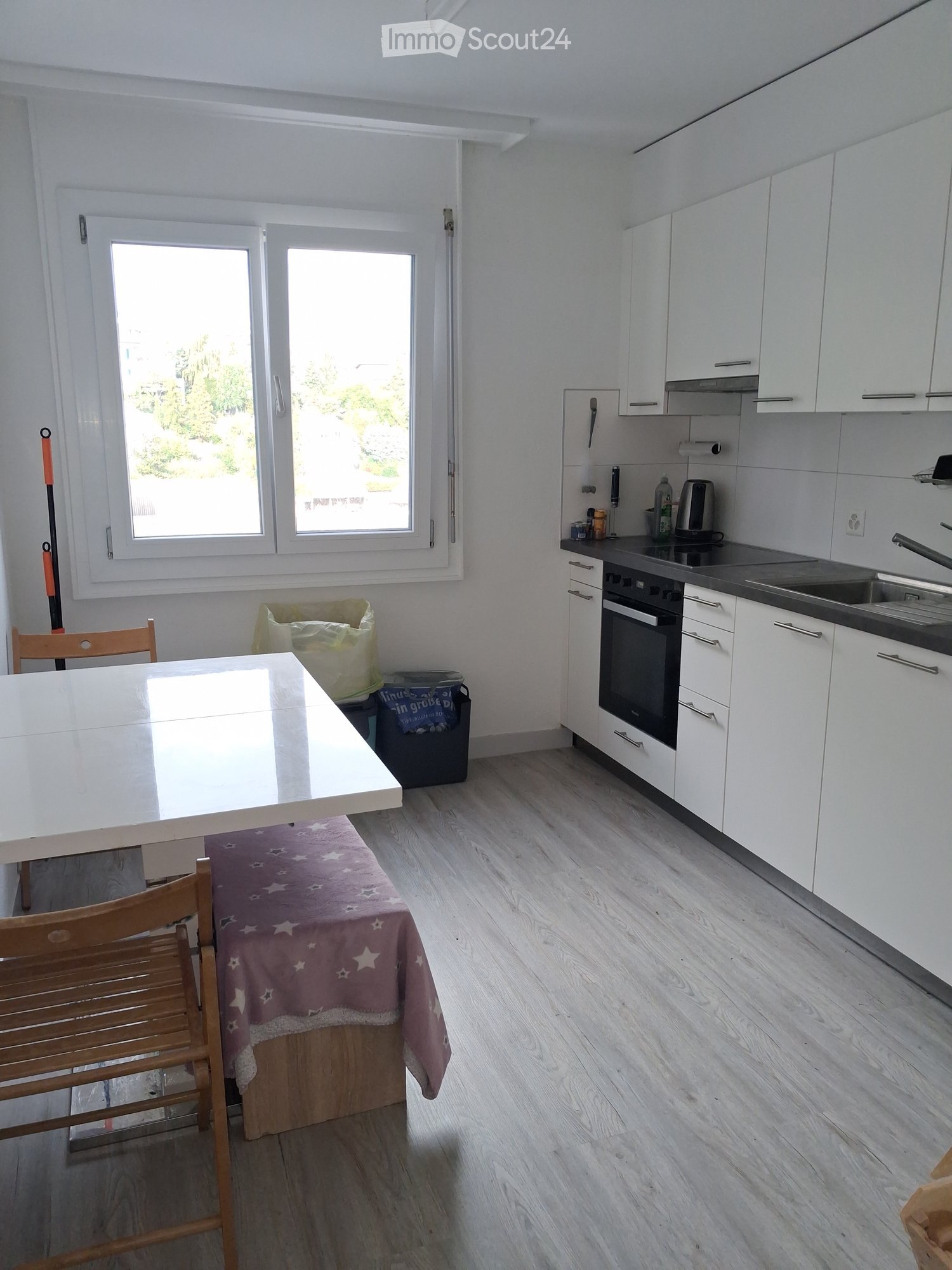 Modern kitchen with white cabinets, wooden flooring, and a dining area featuring a table and chairs. The kitchen includes an oven, sink, and various small appliances.