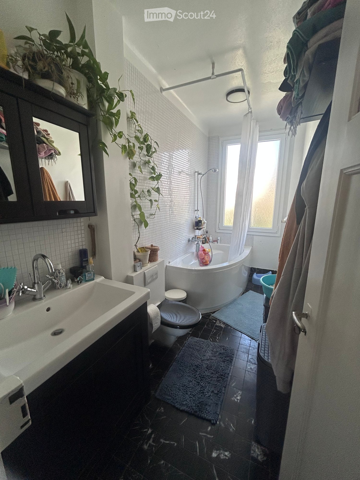 Modern bathroom with a white bathtub, toilet, sink, and mirror. Features a large window, a hanging plant, and a dark tiled floor.