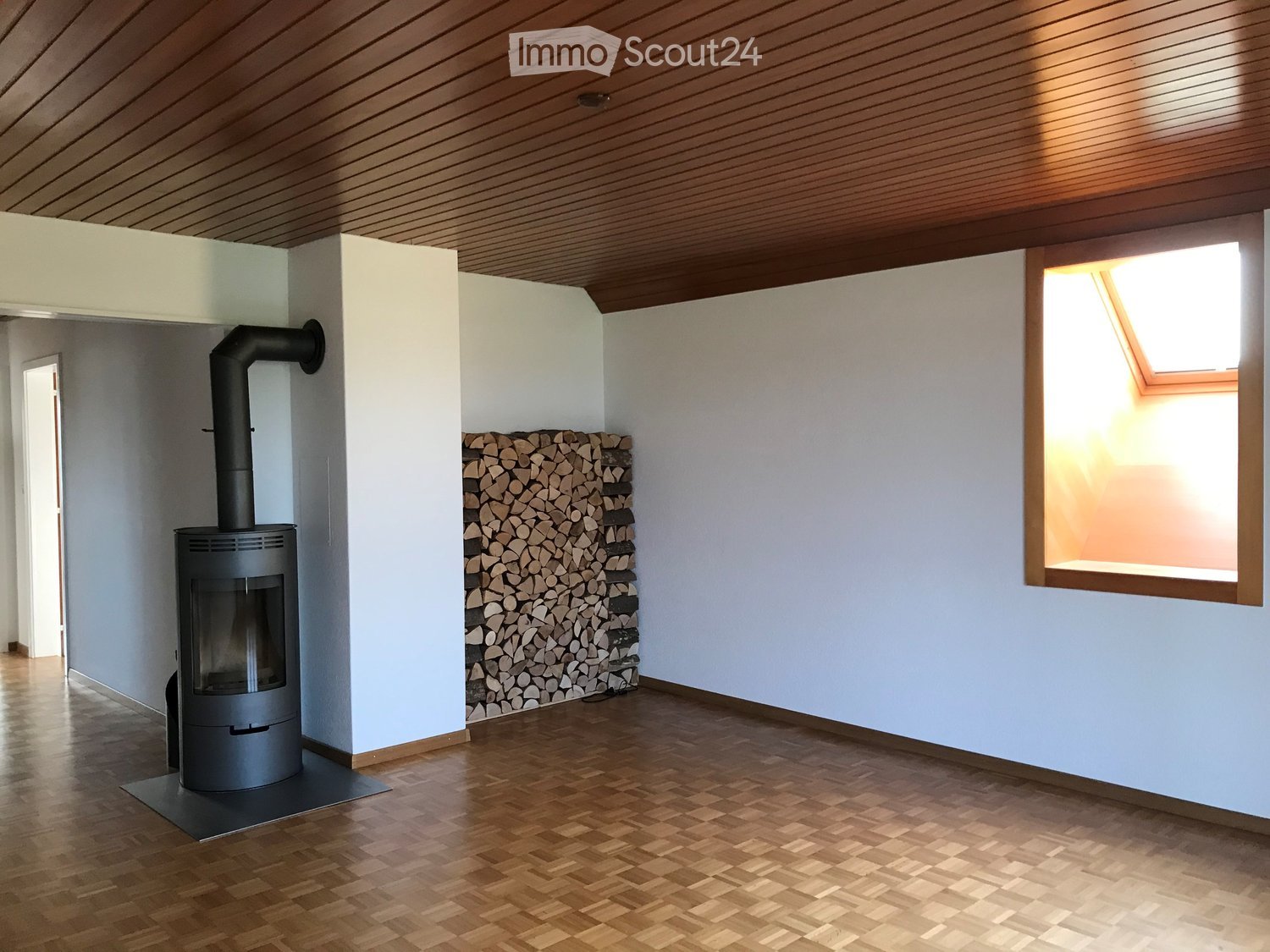Empty room with wooden flooring, white walls, and a modern wood-burning stove with a stack of firewood beside it. A skylight window provides natural light.