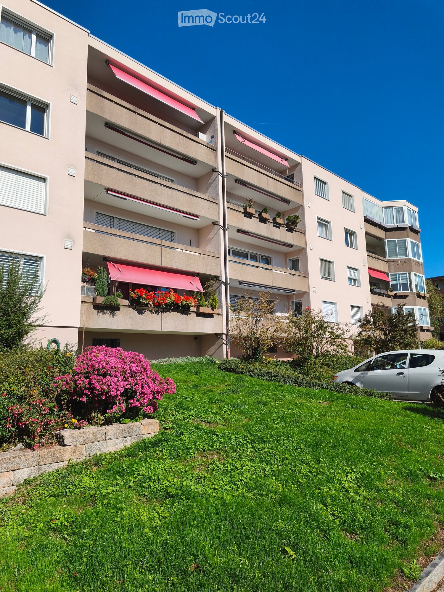 Multi-story residential building with balconies, red awnings, and flower boxes, surrounded by a well-maintained lawn and garden area.